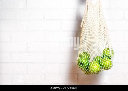 Fresh green apples in eco natural reusable cotton mesh bag. Zero waste concept. White brick background.  Stock Photo