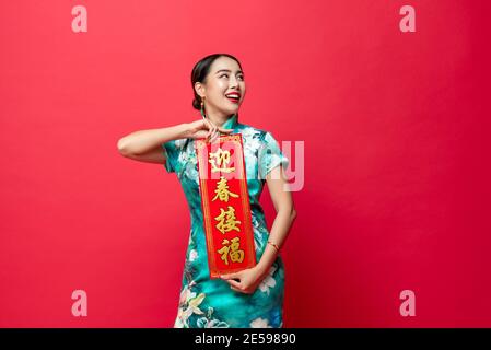 Smiling Asian woman wearing oriental cheongsam dress presenting Chinese new year label and looking upward to copy space in red isolated studio backgro Stock Photo