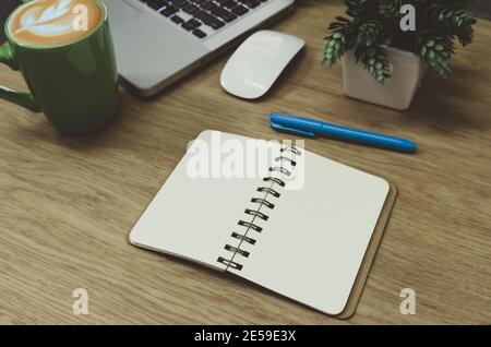 blank notebook lying on a wooden table and a green coffee mug on the computer Stock Photo