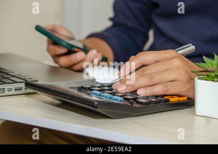 businessman using a calculator holds a pen and a mobile phone on a desk with a laptop working from home. Stock Photo