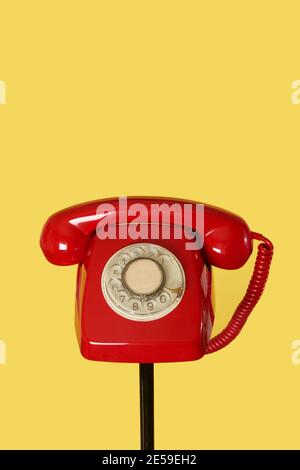 a red landline rotary dial telephone on the top of a black tubular stand, on a yellow background, with some blank space on top Stock Photo