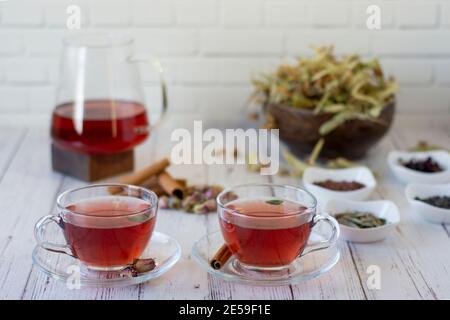 Various herbs brewed in a glass teapot on wood in front of the white wall. Two glasses of linden, pomegranate flowers, green tea, grape seeds, olive l Stock Photo