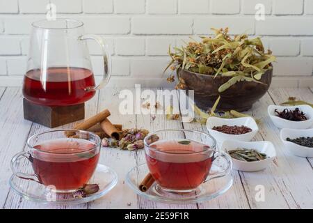 Various herbs brewed in a glass teapot on wood in front of the white wall. Two glasses of linden, pomegranate flowers, green tea, grape seeds, olive l Stock Photo
