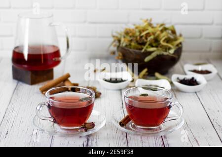 Various herbs brewed in a glass teapot on wood in front of the white wall. Two glasses of linden, pomegranate flowers, green tea, grape seeds, olive l Stock Photo