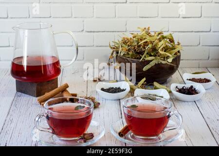 Various herbs brewed in a glass teapot on wood in front of the white wall. Two glasses of linden, pomegranate flowers, green tea, grape seeds, olive l Stock Photo