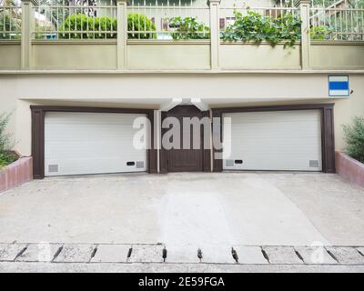 Two symmetrical garages with closed white roller shutters and iron brown door between them Stock Photo
