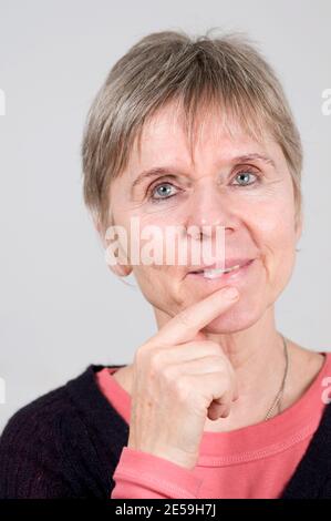 Head portrait of an elderly woman in front of a light gray background. Looking happily at the camera with her left index finger on her chin. Stock Photo