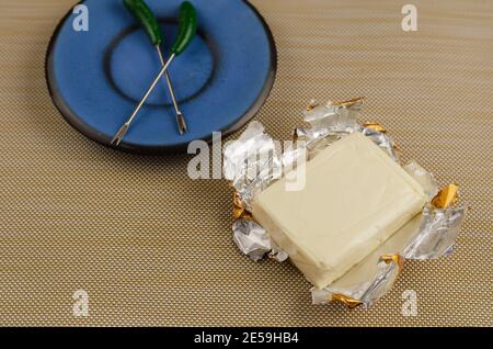Rectangular piece of processed cheese in an open foil package on a brown background. Skewers for stabbing in a blue saucer. Cheese ready to eat. Dairy Stock Photo
