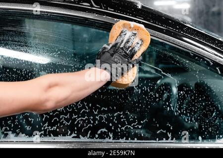 Close up of car wash worker wearing protective gloves and washing car window with soapy sponge. Cleaning services. Stock Photo