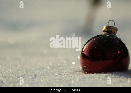 A New Year's red glass ball lies in the sparkling snow outdoors in sun glare on a frosty winter day. Space for text. Christmas card. Stock Photo
