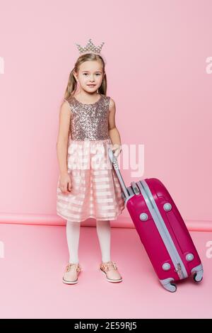 full length of happy little girl in crown and dress standing near baggage on pink Stock Photo