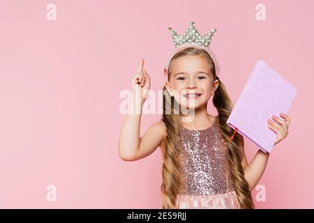 smiling little girl in crown holding book and pointing with finger isolated on pink Stock Photo