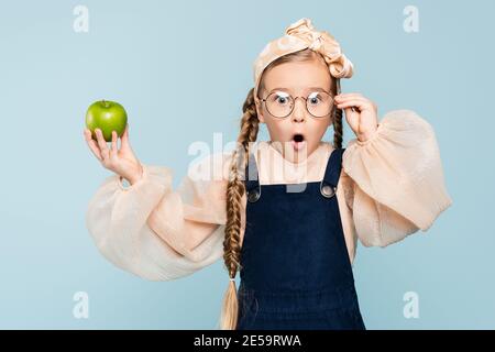 shocked kid adjusting glasses and holding green apple isolated on blue Stock Photo