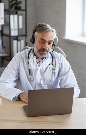 Portrait of senior grey-haired male doctor in his office using laptop for video chat with a patient. Online consultation with doctor for diagnoses and Stock Photo