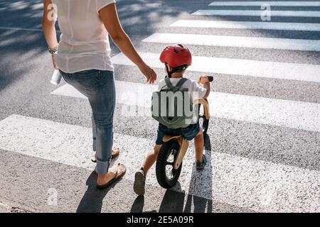 Rear view of a child on a balance bike crossing a road with his mother.