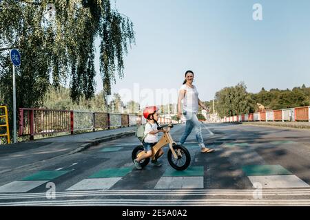 Side view of child on balance bike and his mother crossing road.