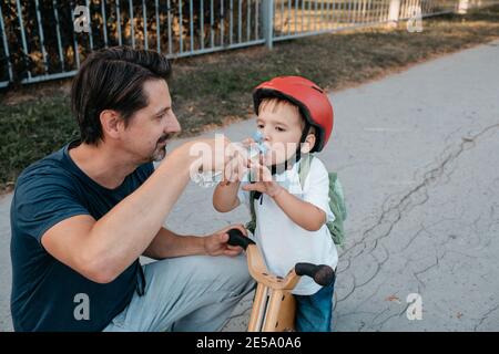 Father offering a bottle of water to his son on a balance bike. Young child on a bicycle taking a break from cycling to have a drink. Stock Photo