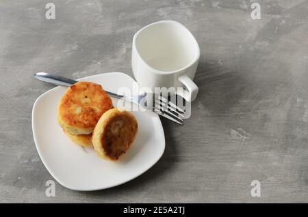 A few pancakes on a white plate with a fork and an empty mug on a textured gray background. Selective focus. Stock Photo