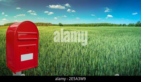 Big red modern postbox with white empty note space for address is standing outdoor in front of beautiful countryside landscape background with farm gr Stock Photo