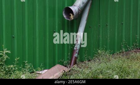 Rainwater flows in jet from gutter pipe with fall leaf inside. Autumn rain pouring from old weathered downspout at rustic backyard with wet grass lawn Stock Photo