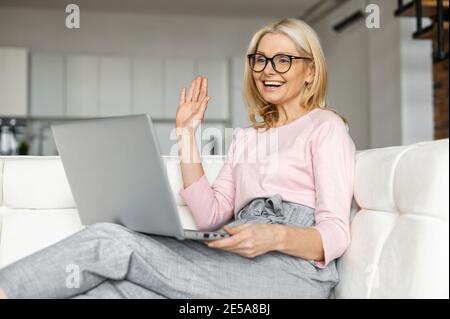 A smiling middle aged woman is sitting in the living room, relaxing and talking to her friends or family via video call at the laptop screen, a glad Stock Photo