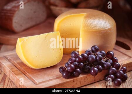 yellow loaf of hard cheese on a chopping board, grapes, still life Stock Photo