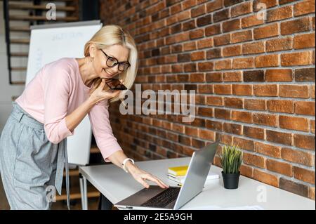 Confident middle aged businesswoman in glasses using voice recognition function on a smartphone. Modern female entrepreneur looking at the laptop Stock Photo