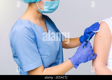 Cropped photo of a medical worker in medical uniform and disposable mask putting covid-19 vaccine on a patient's forearm against a gray background. Stock Photo