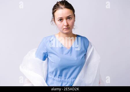A portrait of a tired doctor who takes off her protective gown after a work shift. Pandemic and coronavirus concept on gray background with empty side Stock Photo