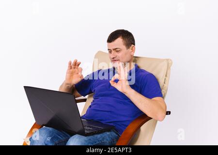 A deaf senior man with a laptop shows a sign to the camera that everything is fine while sitting in a leather chair on a white background. Online non Stock Photo