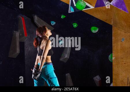 Young woman preparing to climb in bouldering gym, applying chalk on hands and examining route on wall Stock Photo