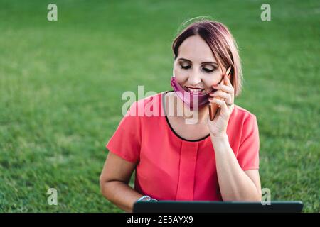 Woman 40 years old working outdoors with laptop during coronavirus outbreak - Smiling entrepreneur sitting on lawn with personal computer while callin Stock Photo