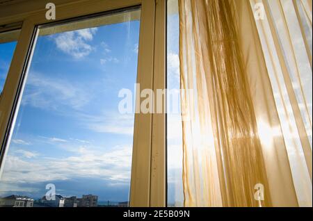 Sun shining through curtains and window. Sunlit apartment on sunset. City view and blue sky. Stock Photo