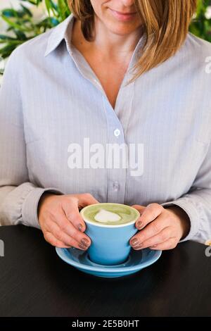 Woman holding blue cup with hot trendy green latte with art heart on the foam on black wooden table background, healthy drink with latte art, morning Stock Photo