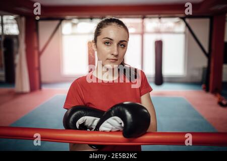 A female athlete wearing boxing gloves after a workout in a gym. A portrait of a young woman sweating after a difficult kickboxing class and looking i Stock Photo
