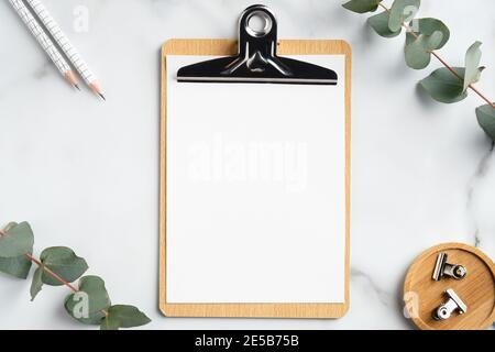 Mockup of wooden clipboard with blank paper on marble table with office supplies and eucalyptus branches. Top view with copy space, flat lay. Stock Photo