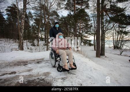 A man looks after a woman in a wheelchair. They are out for a walk. Winter evening. They are wearing medical masks. High quality photo Stock Photo