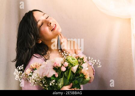 attractive young woman enjoying and feeling the sun holding a flowers bouquet with simple pink dress. Smiling and confident woman during spring. Stock Photo