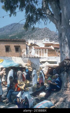 1984 Morocco Chefchaouen - Chefchaouen, Shafshāwan, also Chaouen, is a city in northwest Morocco. It is noted for its buildings in shades of blue. Chefchaouen is situated just inland from Tangier. The beauty of Chefchaouen's mountainous surroundings are enhanced by the contrast of the brightly blue painted medina (old town). Berber women in traditional hat and tourists in the Old town of Chefchaouen Morocco North Africa Stock Photo