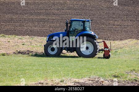 blue agricultural tractor without driver stands on a green meadow in front of a plowed field in the background, sunshine, during the day Stock Photo