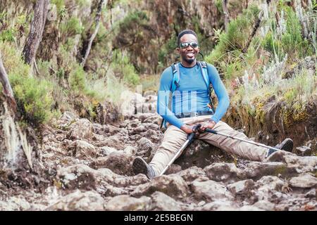 Portrait of a cheerfully smiling African-American Ethnicity young man in sunglasses sitting with backpack and trekking poles and resting on mountain t Stock Photo