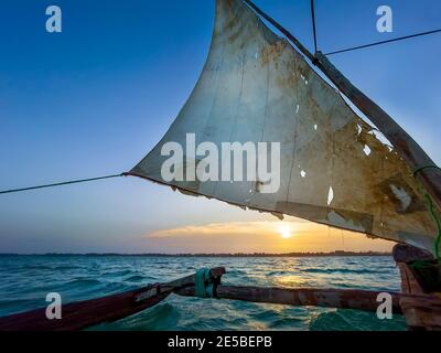 Old traditional maritime traditional vessel Dhow boat sailing under torned sail in the open Indian ocean near Zanzibar island in beautiful sunset, Tan Stock Photo