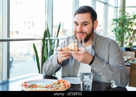 Happy smiling caucasian man eating Italian pizza in pizzeria keeps social distance. Delicious pizza in cafe. Stock Photo