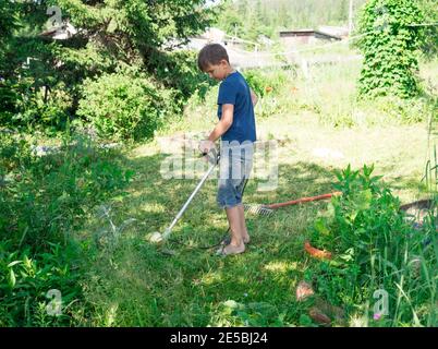 Boy, 11 years old, mows the grass with an electric scythe on the lawn in the yard of a house on a sunny summer day. Stock Photo
