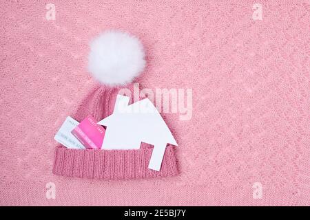 winter flat lay with credit cards, paper house and a hat with pompom on pink background.