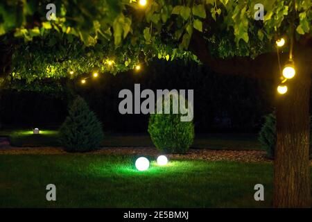illumination park light garden with electric ground ball lantern with stone mulch and thuja bushes in outdoor landscaped park with garland of warm lig Stock Photo
