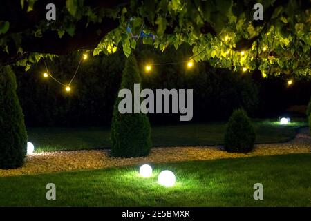 illumination park light garden with electric ground ball lantern with stone mulch and thuja bushes in outdoor landscaped park with garland of warm lig Stock Photo