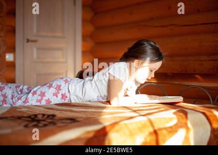 close-up a girl reading a book lies on her stomach in her pajamas on the bed in the bedroom of a log cabin in the village Stock Photo