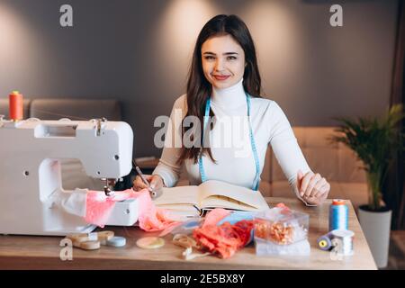 Young dressmaker woman sews clothes on sewing machine. Pretty and beautiful seamstress in workshop. Creating online clothing design courses. Stock Photo