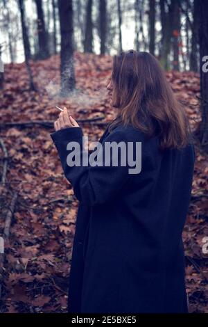 young brunette woman smoking cigarette in the forest in autumn Stock Photo